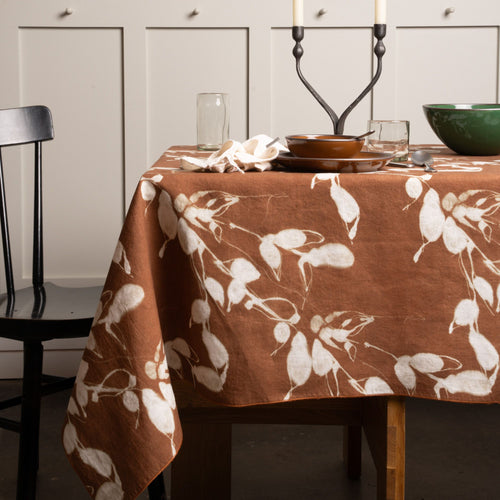 Brown tablecloth with white leaf pattern, styled on a dining table with dinnerware, glassware, and candles.