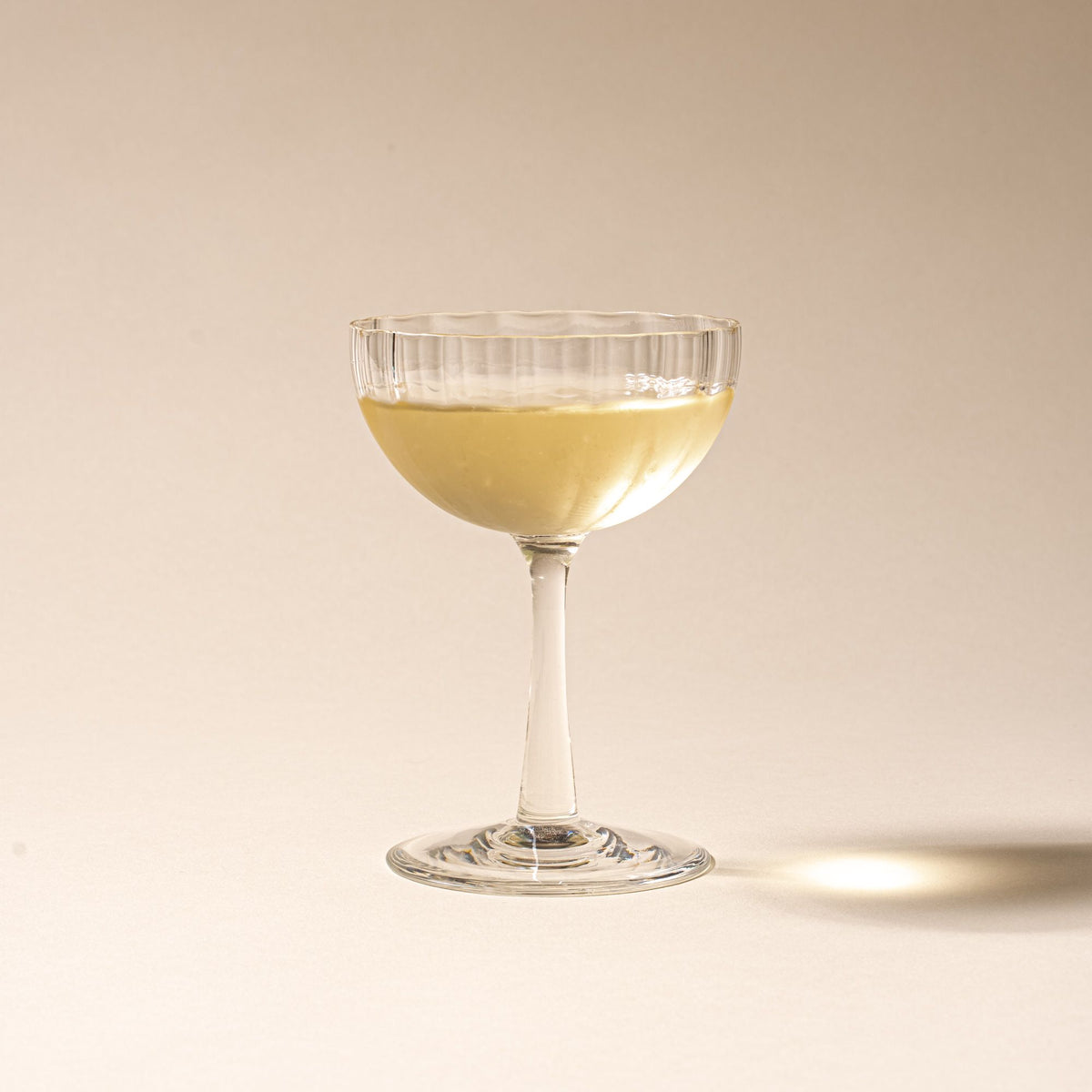 White wine in a clear ribbed glass coupe with a tall stem casting a soft shadow on a beige background.