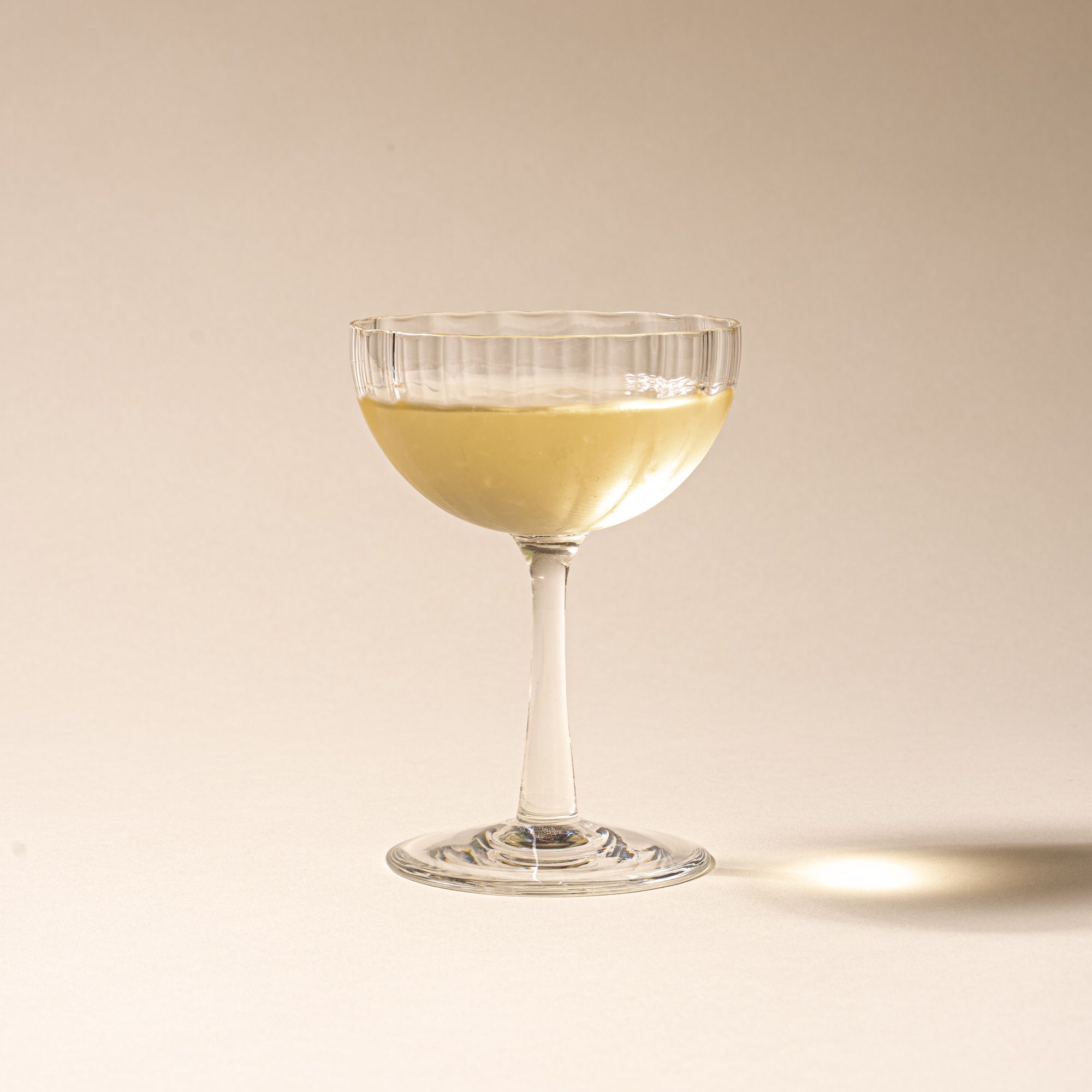 White wine in a clear ribbed glass coupe with a tall stem casting a soft shadow on a beige background.