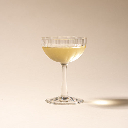 White wine in a clear ribbed glass coupe with a tall stem casting a soft shadow on a beige background.