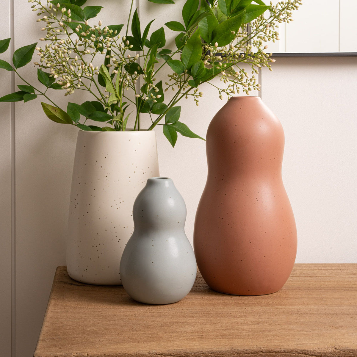 Three ceramic vases in cream, blue-gray, and terracotta sit on a wooden surface; the cream vase is filled with lush green leafy branches with small white buds