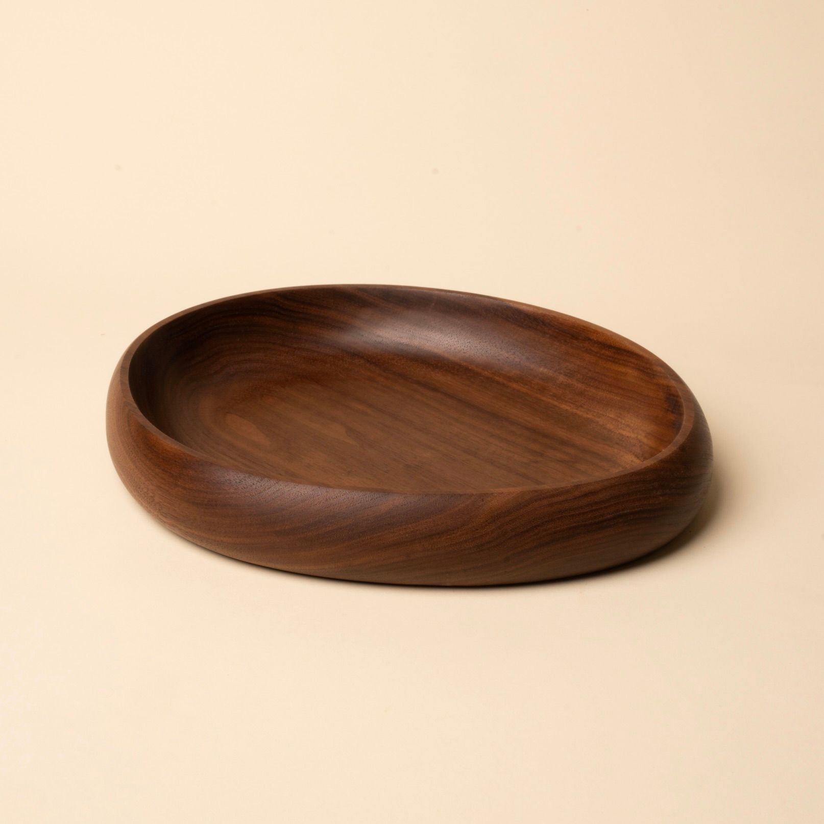 Shallow carved round walnut wood dish with smooth curved edges, resting on a beige background.