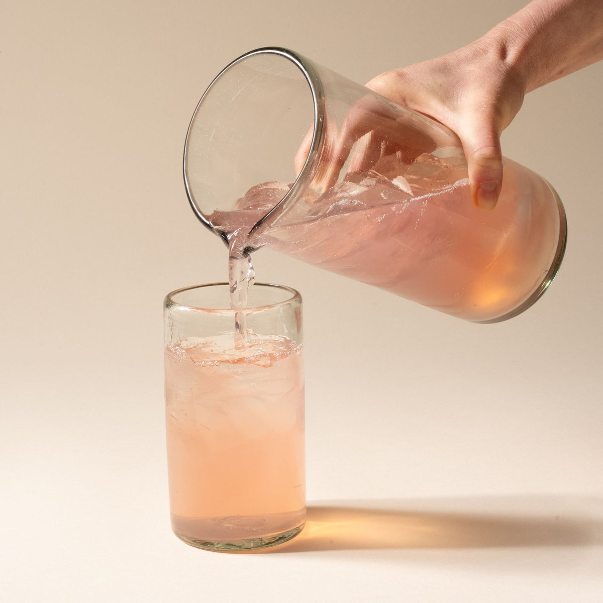 A hand holds a clear glass pitcher with small spout at the top, and pours a drink into a tall tumbler.