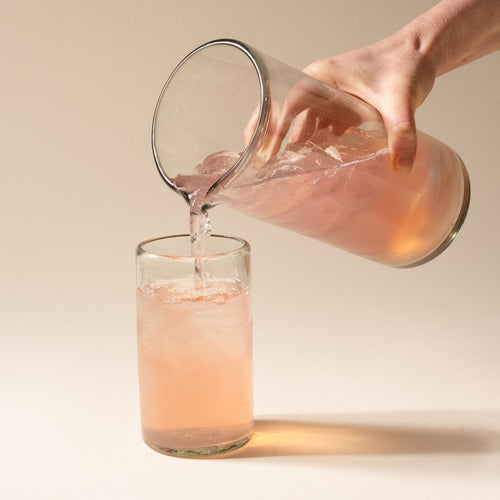 A hand holds a clear glass pitcher with small spout at the top, and pours a drink into a tall tumbler.
