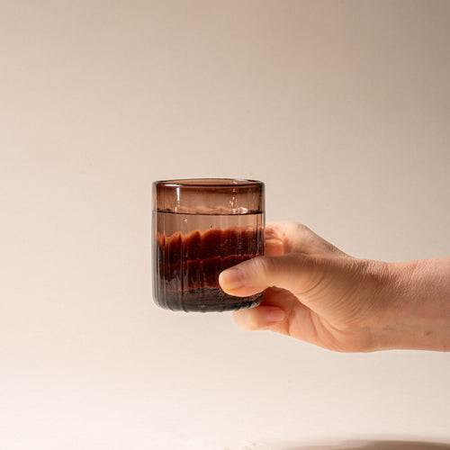 A hand holds an amber-brown glass tumbler with vertical ridges, filled with water, set against a neutral background.