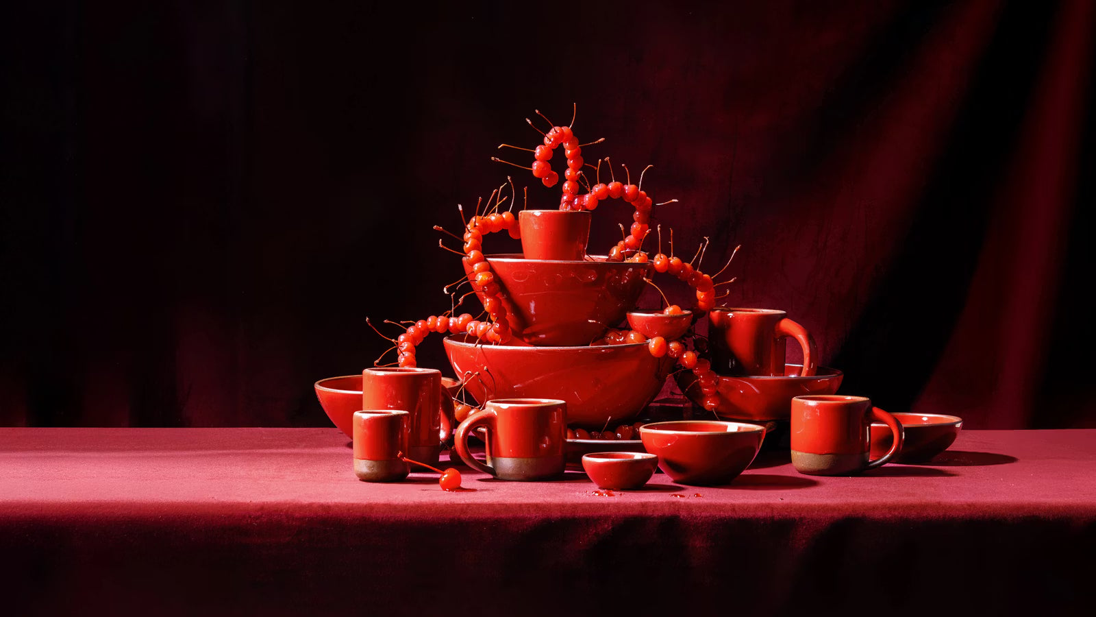 Glossy red ceramic cups, mugs and bowls stacked into a tower, draped with glossy maraschino cherries against a deep red backdrop.