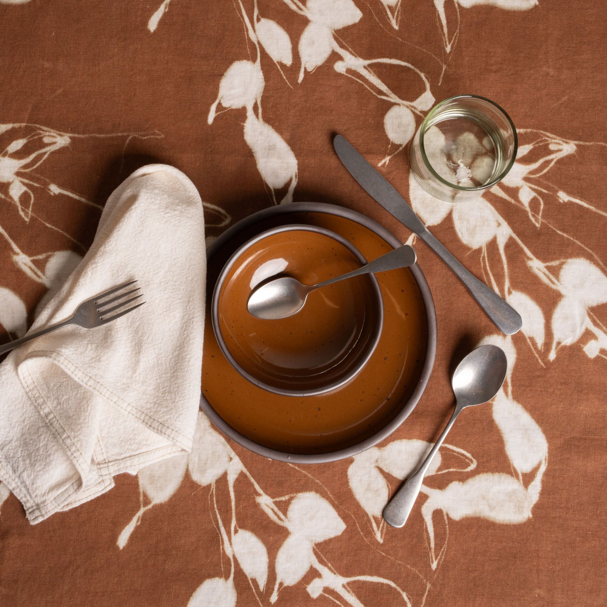 Place setting on brown floral tablecloth with ceramic dishes, silverware, glass, and a folded cloth napkin.