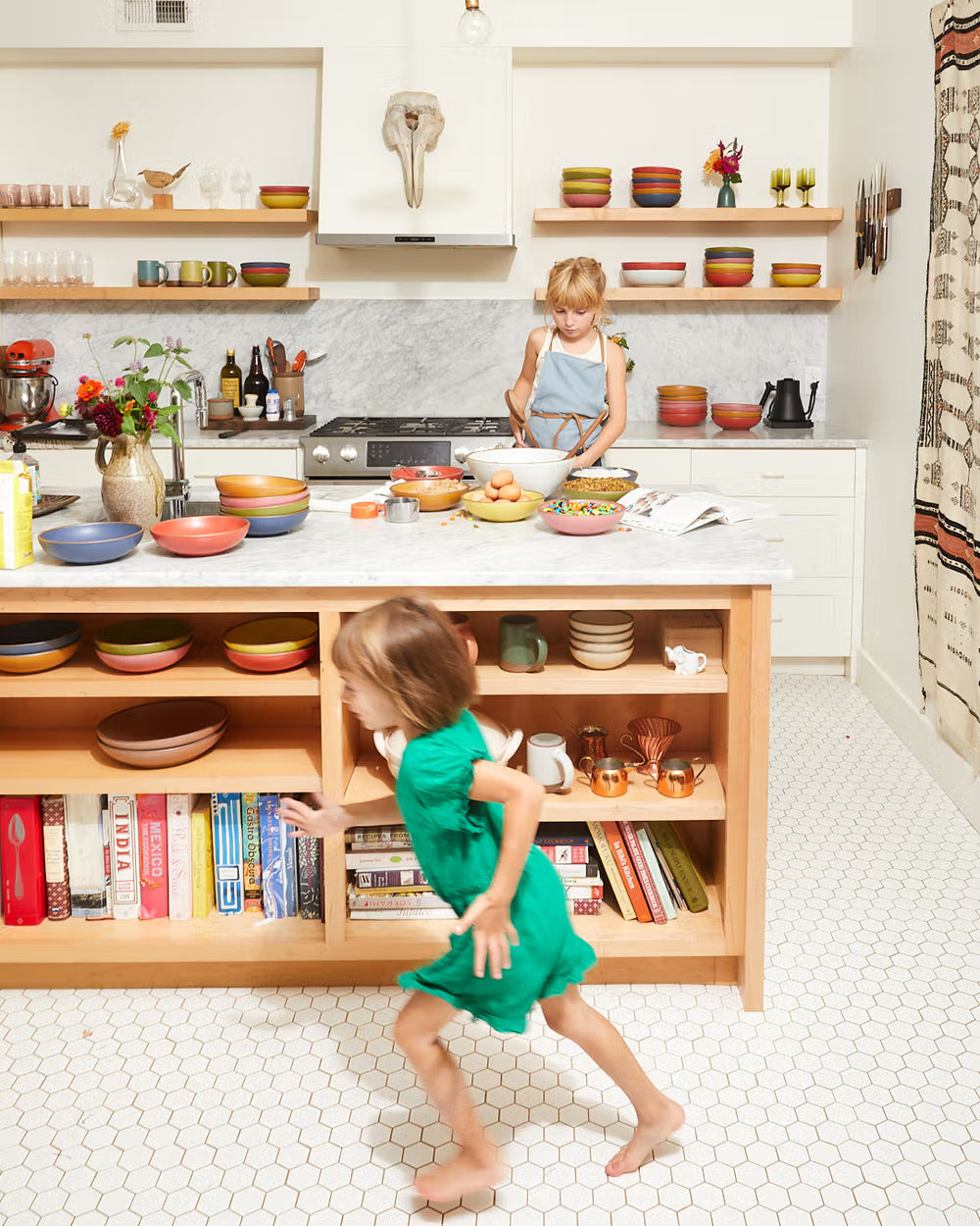A child wearing a blue apron is mixing ingredients in a bowl on a marble kitchen island filled with colorful ceramic dishes. Another child in a green dress runs past shelves stocked with books and more ceramics, set against a backdrop of an eclectic, cozy kitchen.