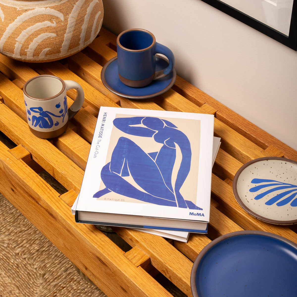 A Matisse art book sits on a wooden bench beside blue and cream ceramics, including mugs and plates with abstract patterns inspired by the artist’s cut-outs.