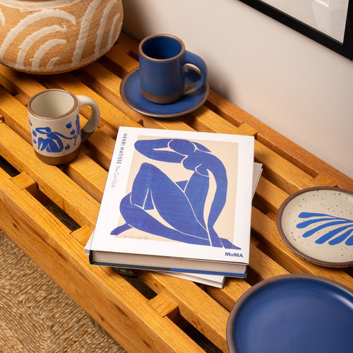 A Matisse art book sits on a wooden bench beside blue and cream ceramics, including mugs and plates with abstract patterns inspired by the artist’s cut-outs.