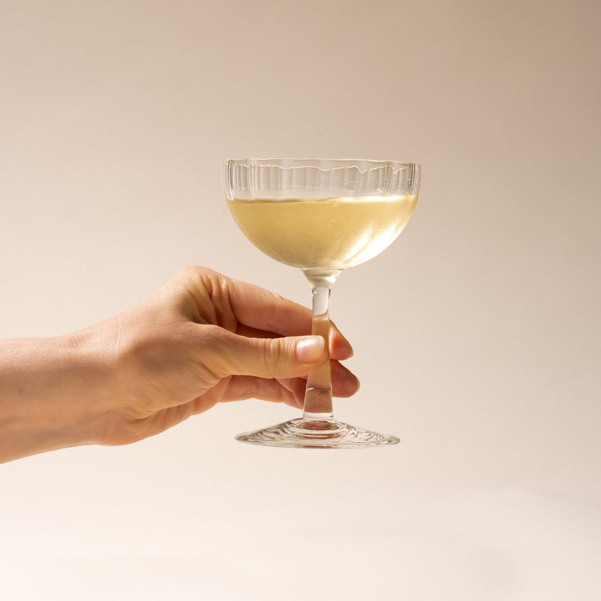 A hand holds out white wine in a clear ribbed glass coupe with a tall stem casting a soft shadow on a beige background.