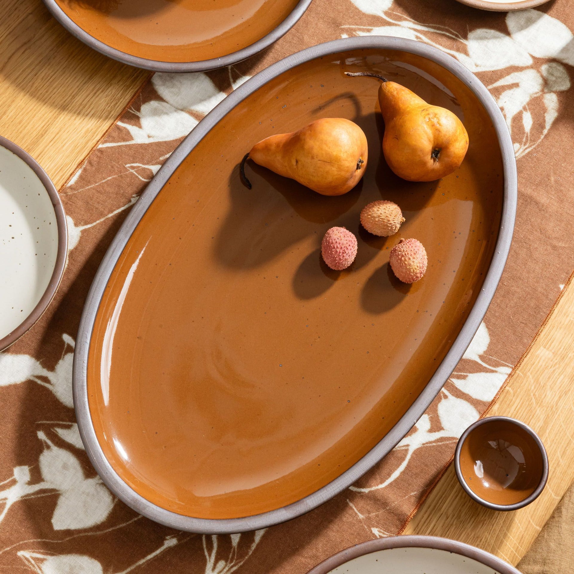 Glossy caramel brown oval platter with two pears and three lychees on a patterned table runner with matching dishes.