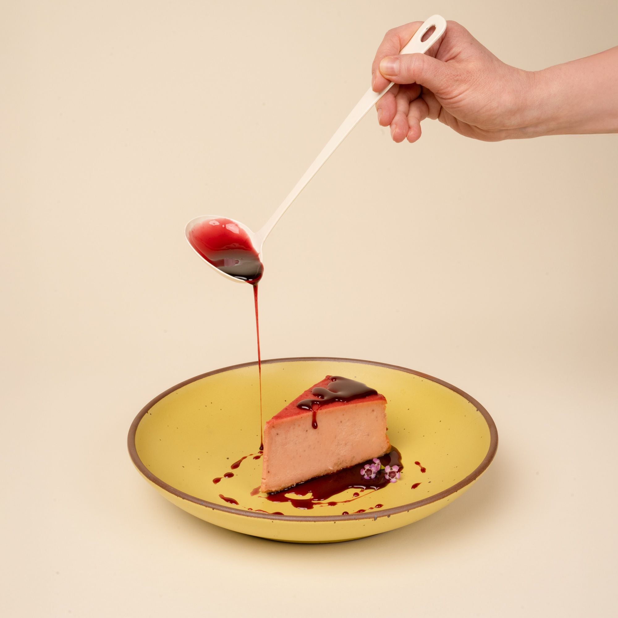 A hand holds a white enamel sauce ladle filled with a red glaze and pours it over a slice of cheesecake on a yellow bowl.
