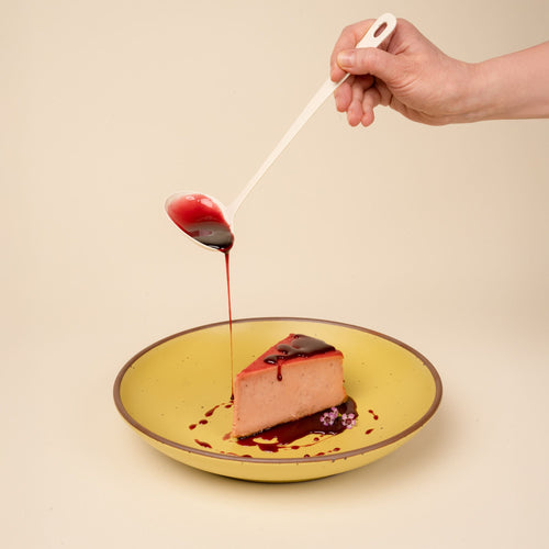 A hand holds a white enamel sauce ladle filled with a red glaze and pours it over a slice of cheesecake on a yellow bowl.