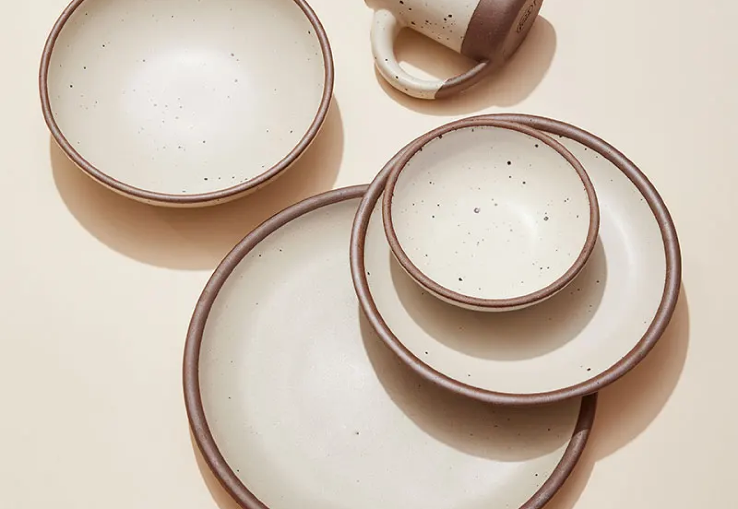 A 7-piece dinner setting of ceramic plates and bowls in a warm, tan-toned, off-white color featuring iron speckles.