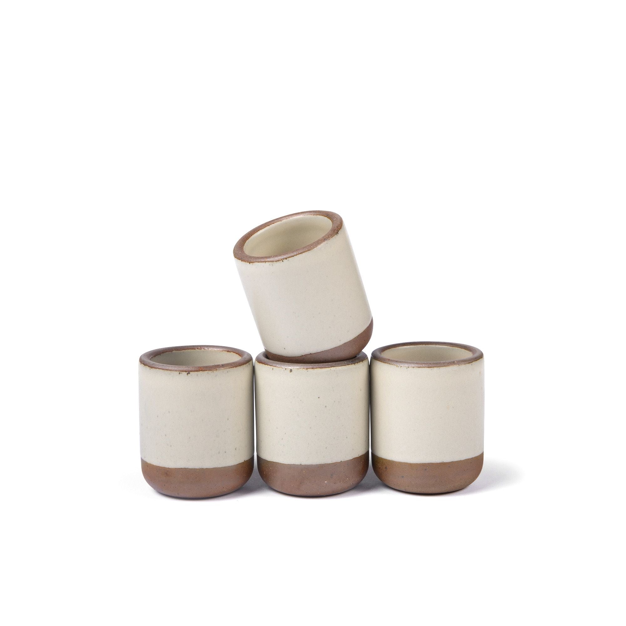 A set of 4 small, short ceramic mug cups in a warm, tan-toned, off-white color featuring iron speckles and unglazed rim and bottom base, stacked in a row of 3, with the 4th on top.