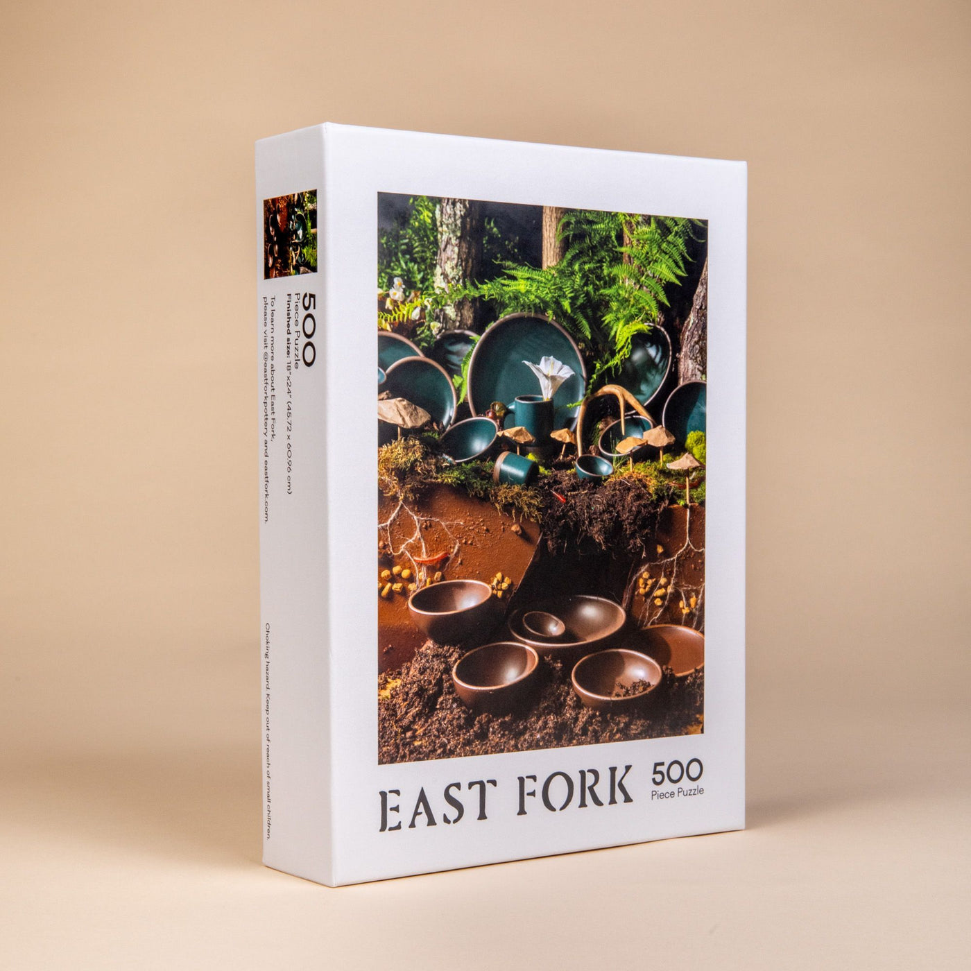 Angled side view of a box of a 500-piece jigsaw puzzle that when put together shows ceramic bowls, plates and mugs in dark teal and brown colors in a natural forest setting.