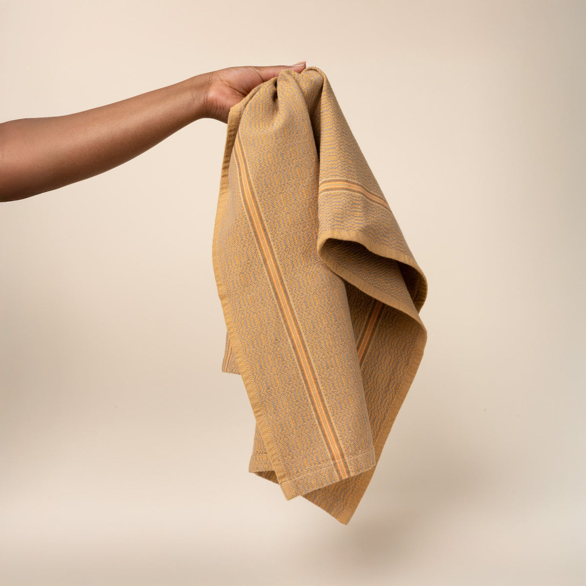 Hand holds out a muted gold woven kitchen towel with a subtle textured pattern and slim striped accent, folded neatly on a clean neutral background.