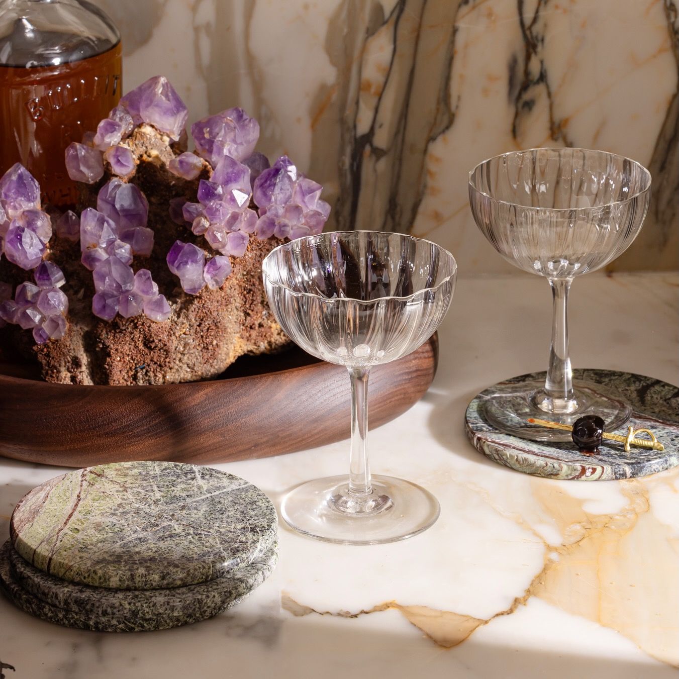 Two coupe glasses on green marble coasters beside amethyst crystals on a wooden tray, set on a marble surface.