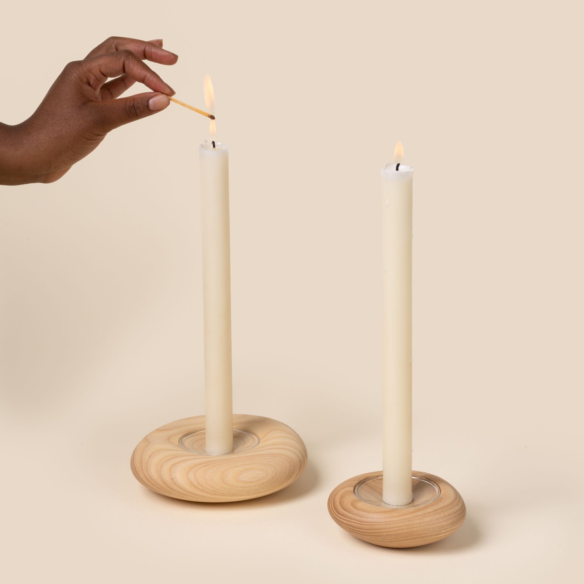 A hand lighting two lit white taper candles in round, light wood candle holders with visible grain, displayed against a neutral background.