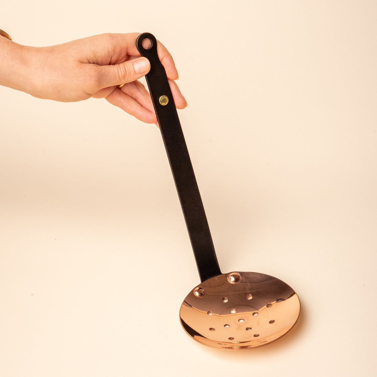 A hand holds up a round copper skimmer with perforated bowl and long black handle featuring a brass rivet and hanging loop