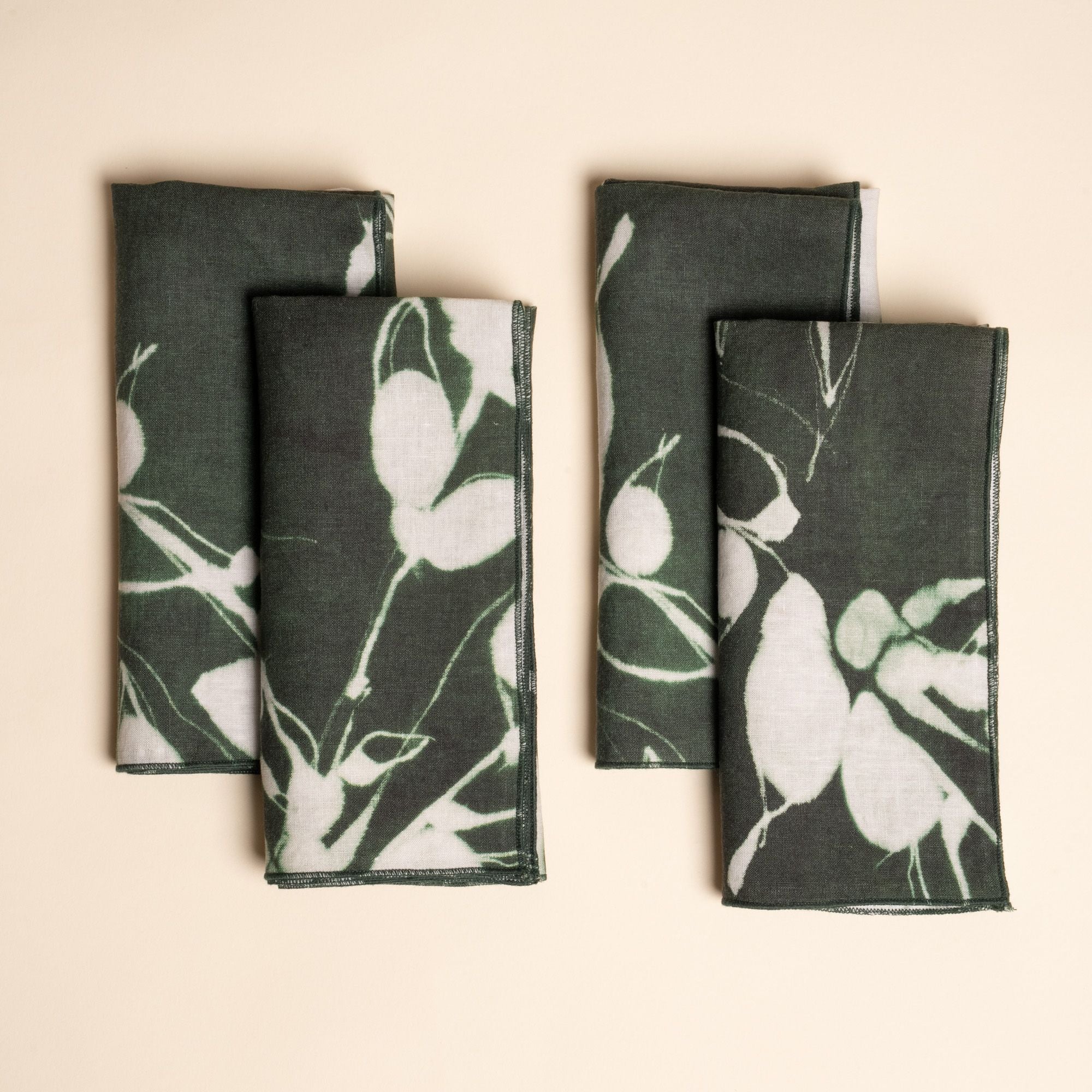 Set of four dark green napkins with white botanical print, neatly folded on a neutral background.
