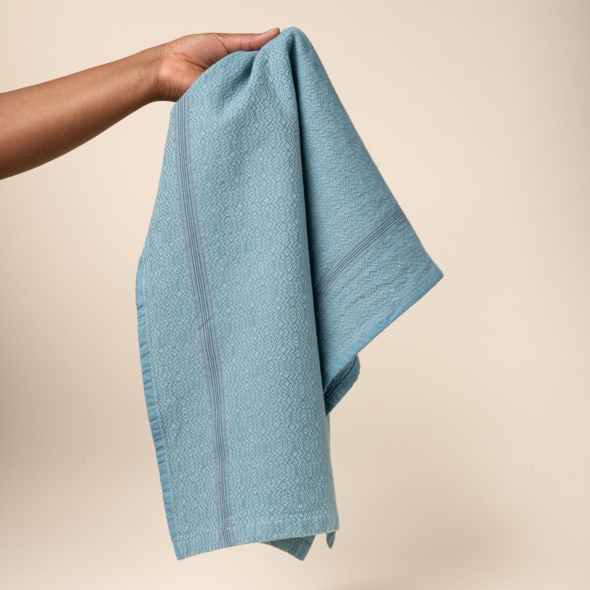 Hand holds out a blue woven kitchen towel with a subtle textured pattern and slim striped accent, folded neatly on a clean neutral background.