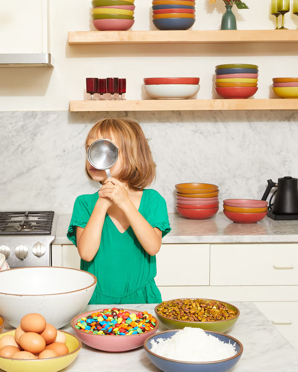 A child in a green dress playfully holds a metal sifter to their face in a kitchen. In front are bowls filled with eggs, colorful candies, nuts, and shredded coconut, with stacked ceramic bowls on the shelves behind.