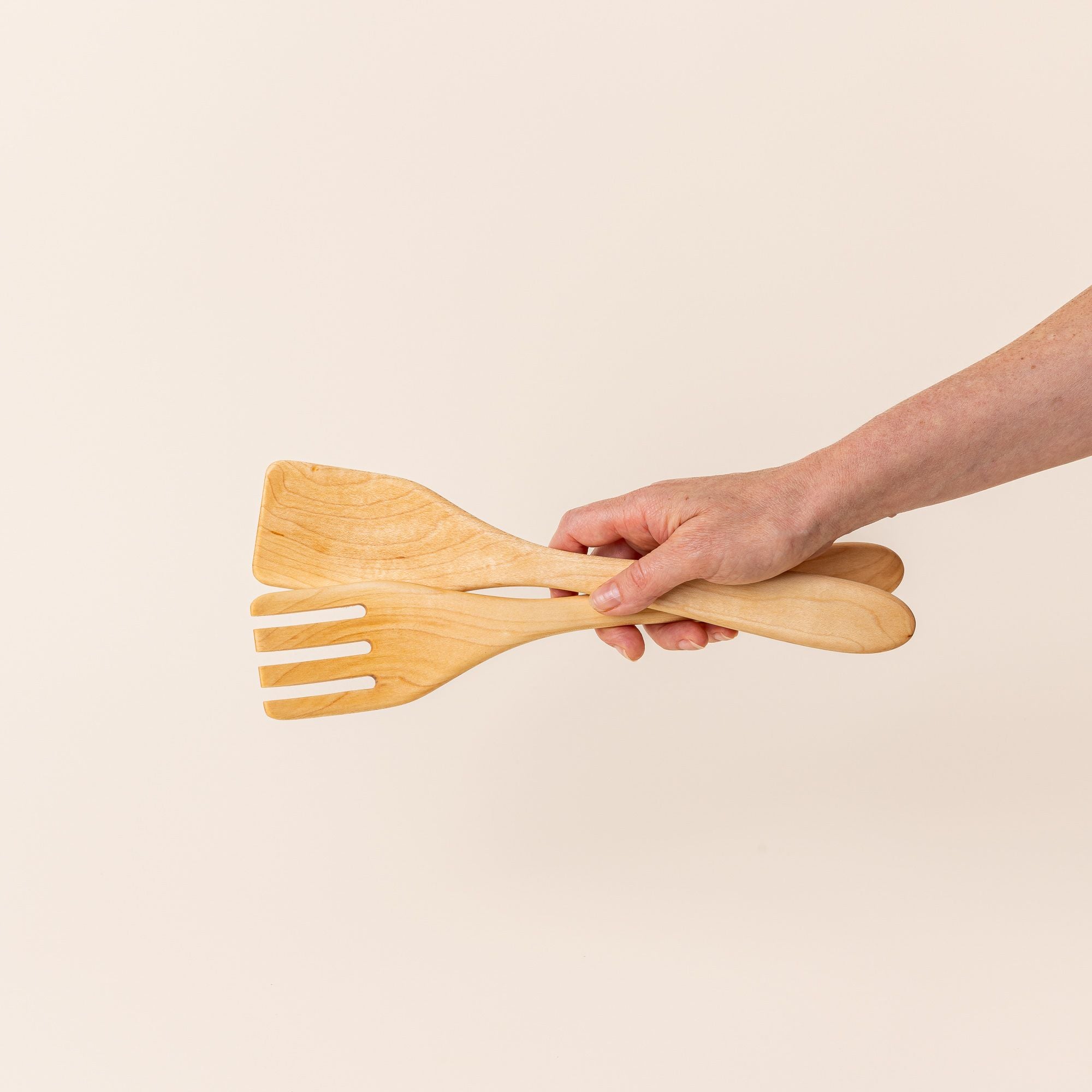 Hand holding a pair of long maple wood serving utensils.
