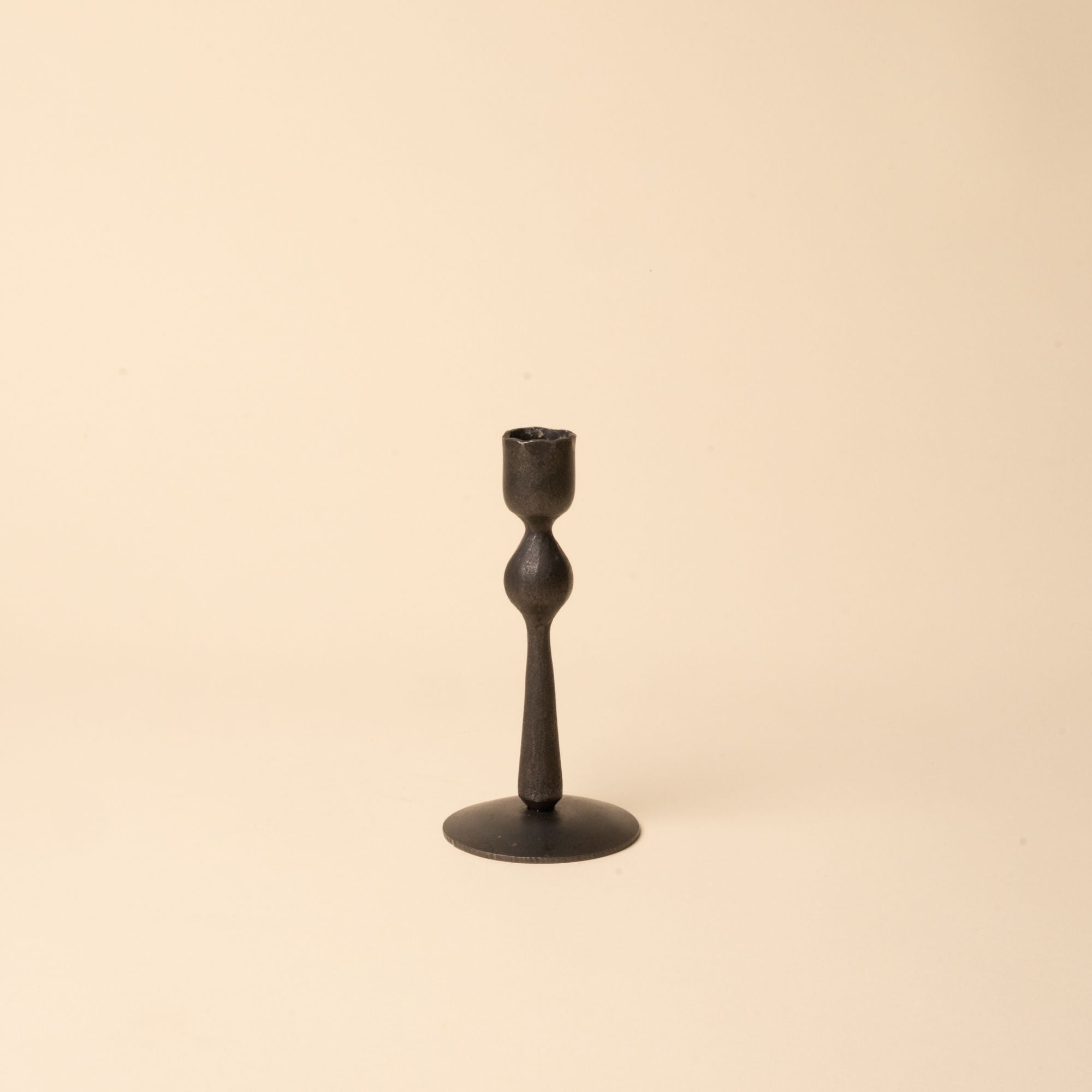 Short minimal black forged iron candlestick with a sculptural shape on a neutral beige background.