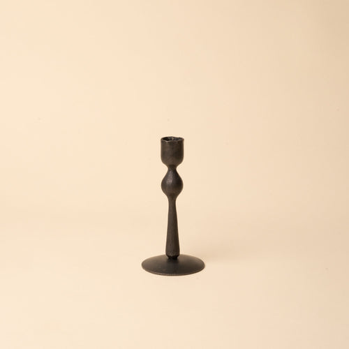Short minimal black forged iron candlestick with a sculptural shape on a neutral beige background.