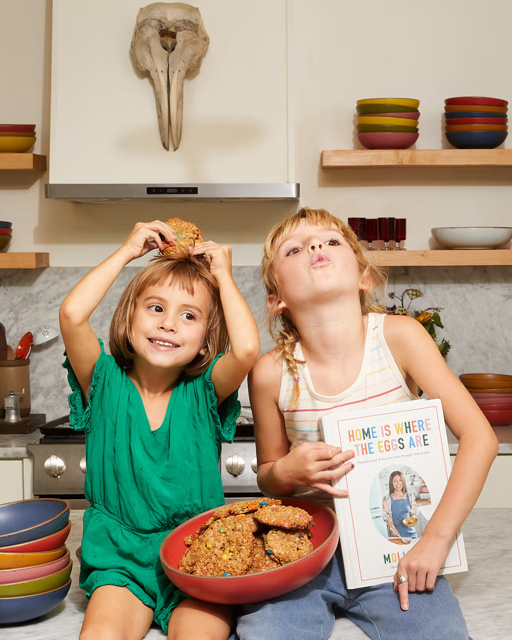 Two children sit in a kitchen with one holding a red bowl of cookies and a cookbook titled "Home is Where the Eggs Are". The other child balances a cookie on their head, with stacked colorful bowls in the background.