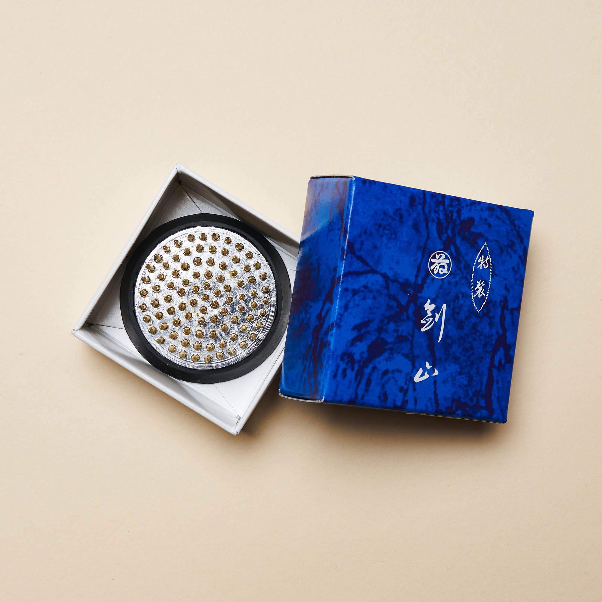 A small "kenzan" – a pin frog made of brass spikes on a flat circular base to assist in flower arrangements sitting inside a white box with a blue top on the right