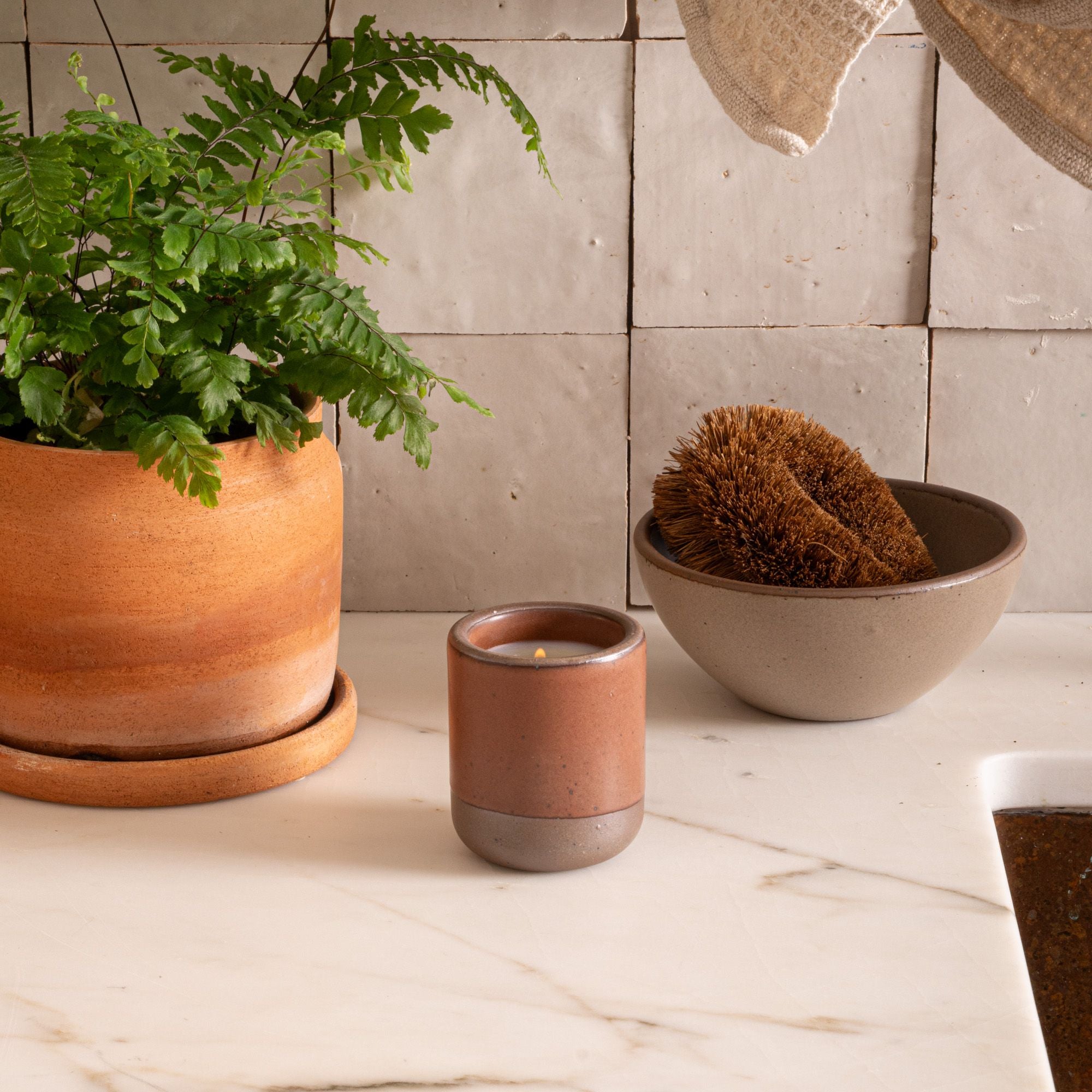 Small ceramic vessel in a cool terracotta color with candle inside on a countertop with a potted plant and dish scrub.