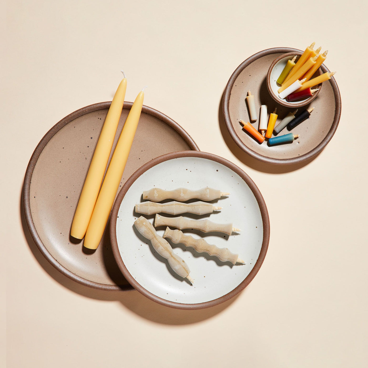 A collection of stoneware plates in varied glazes, one holding two taper candles, another with three wavy candles, and the smallest with assorted colorful candles arranged alongside a vessel on a light neutral background.