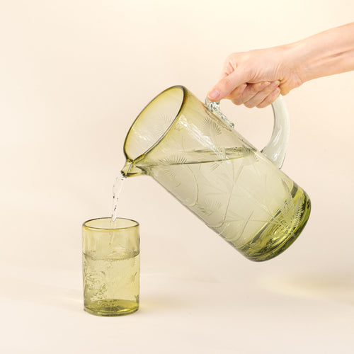 A hand holding a large glass olive pitcher with handle made of recycled glass with etched thistle designs on it, filled with water, and pouring into a matching tumbler.