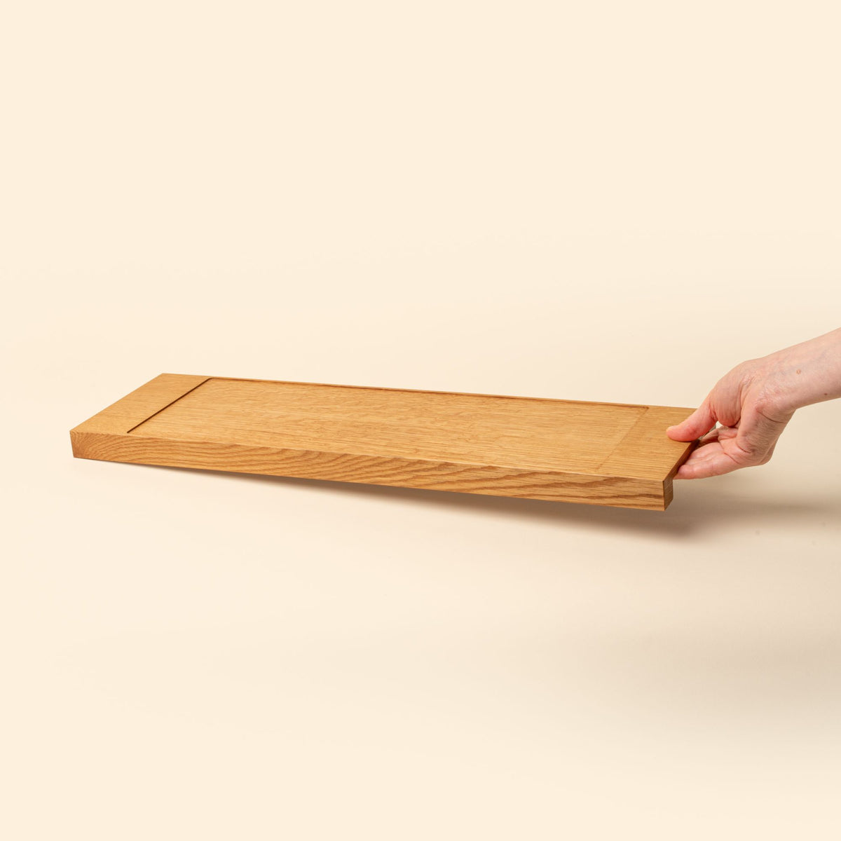 A hand holds out a narrow rectangle of light oak wood with a vertical groove at either end, by the beveled handle on the side.