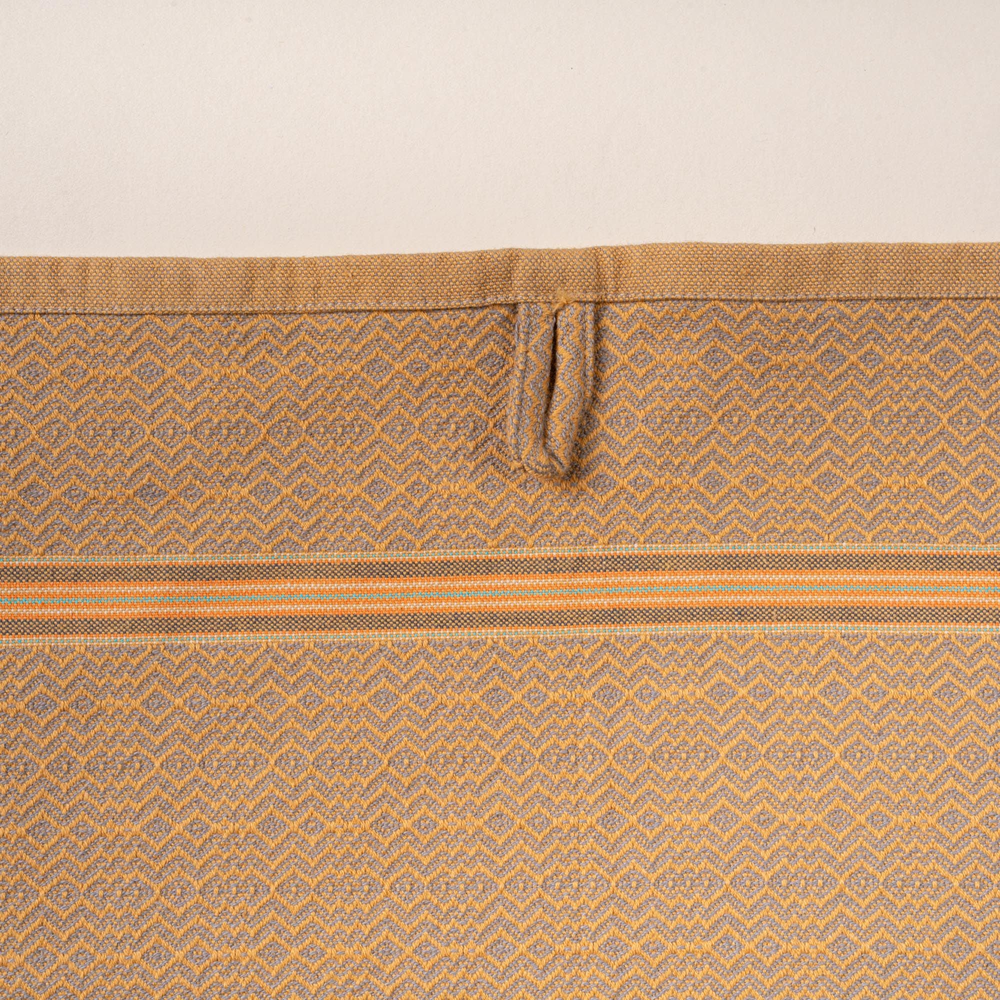 Close-up of a muted gold woven towel with a zigzag-and-diamond texture, stitched hanging loop, and thin striped band detail.