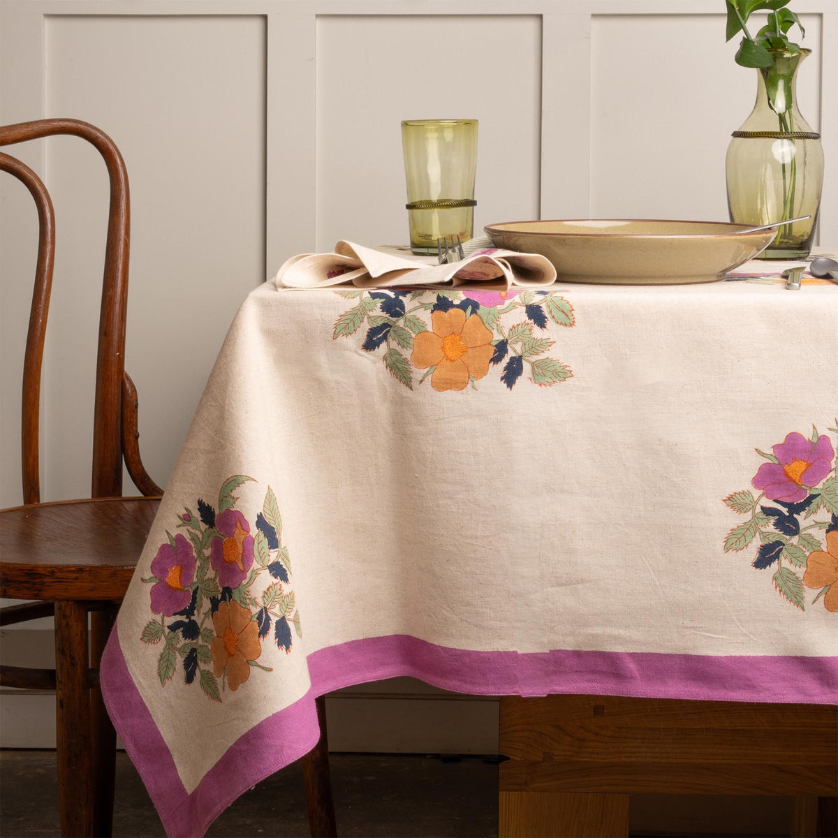 Table set with a cream linen tablecloth with a purple border and floral motifs in purple and orange with green leaves, grey-green large dish and olive green glassware and vases.
