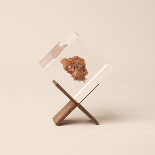 A clear resin cube with a brown mineral inside rests on a small wooden X-shaped stand against a neutral background.