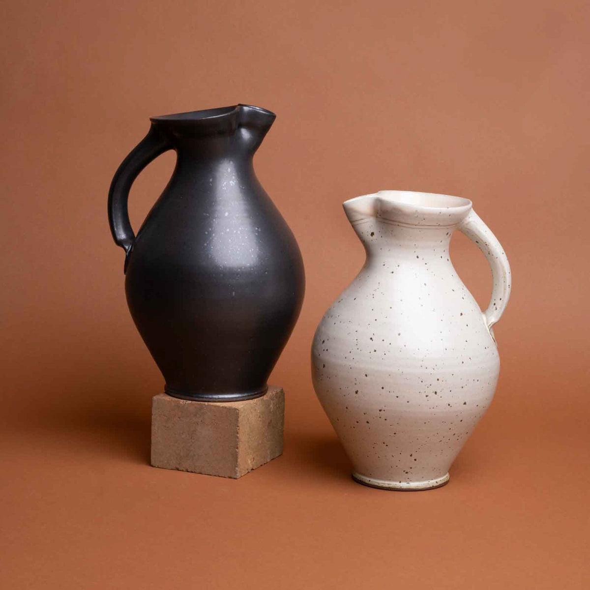 Two ceramic pitchers, one black and one cream with speckles, set against a warm brown background.
