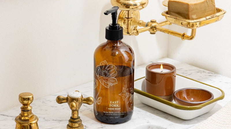 Amber hand-soap bottle on a marble sink beside brass fixtures, a cool terracotta ceramic candle, and a tray with small terracotta dishes in a bright bathroom.
