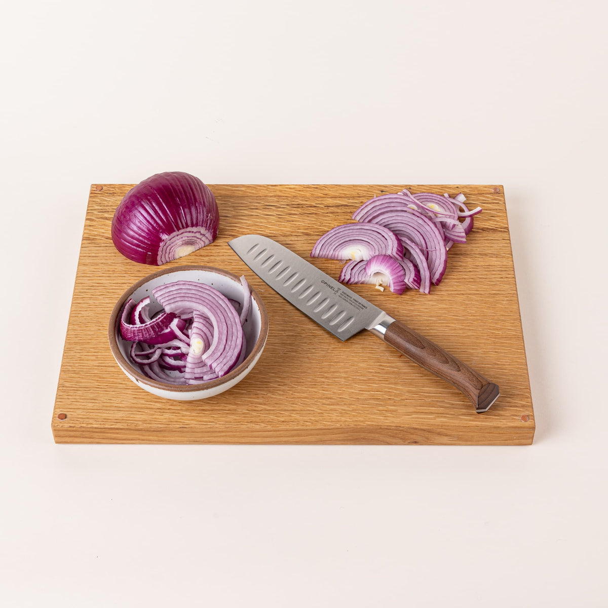 On a cutting board sits a santoku knife with a sharp steel blade with dimples and a beveled dark wood handle with chopped onions