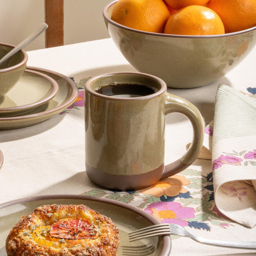 Large stoneware mug with black coffee, citrus bowl, and pastry on a floral tablecloth.