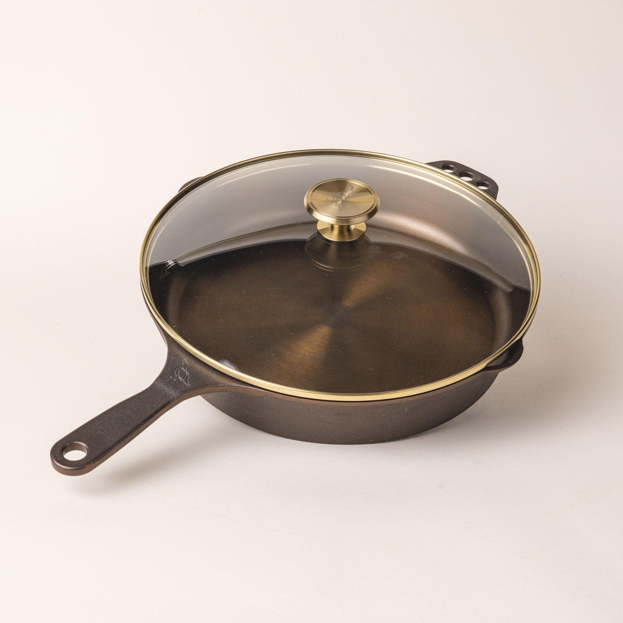 A glass skillet lid with a brass lip and brass handle knob over a cast iron skillet