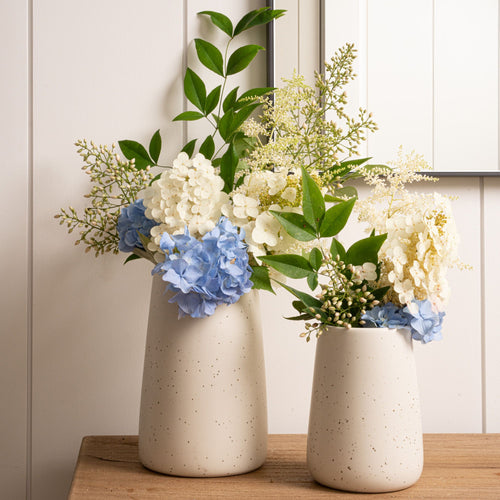 Two cream vases with white and blue hydrangeas and leafy greens on a wooden table near a mirror.