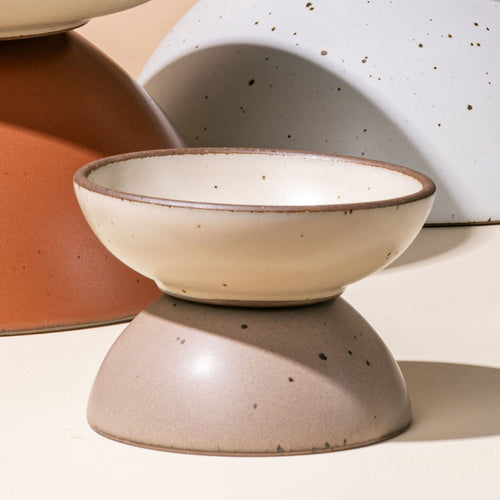 A shallow small ceramic bowl in an off-white color sits on an upside down small potter's bowl in a muted tan color.