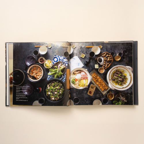 Open cookbook showing a Japanese-style dinner spread with bowls, plates, and shared dishes arranged on a dark tabletop.