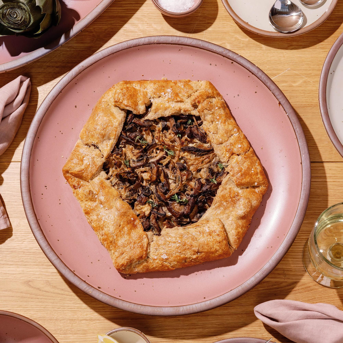 Rustic mushroom galette on a large dusty mauve pink ceramic platter, set on a wooden table with napkins and glassware nearby.