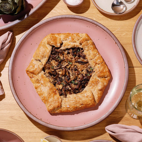 Rustic mushroom galette on a large dusty mauve pink ceramic platter, set on a wooden table with napkins and glassware nearby.